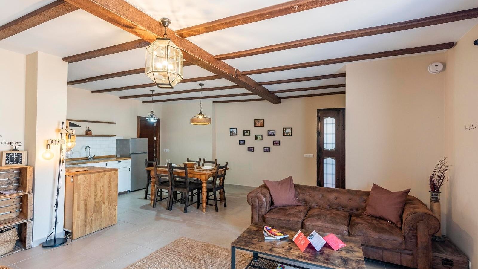 A cozy living room with a brown leather sofa, wooden dining table, and pendant lights. A kitchen is visible in the background with a wooden ceiling.