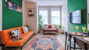 A modern living room with a green accent wall, a sofa, a dining table with chairs, and a large window with curtains. - Modern 4-Bedroom in Manchester (Salford)