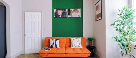 Una habitación con una pared verde decorativa, una puerta blanca, un sofá naranja, un tapete estampado y una planta de bambú.