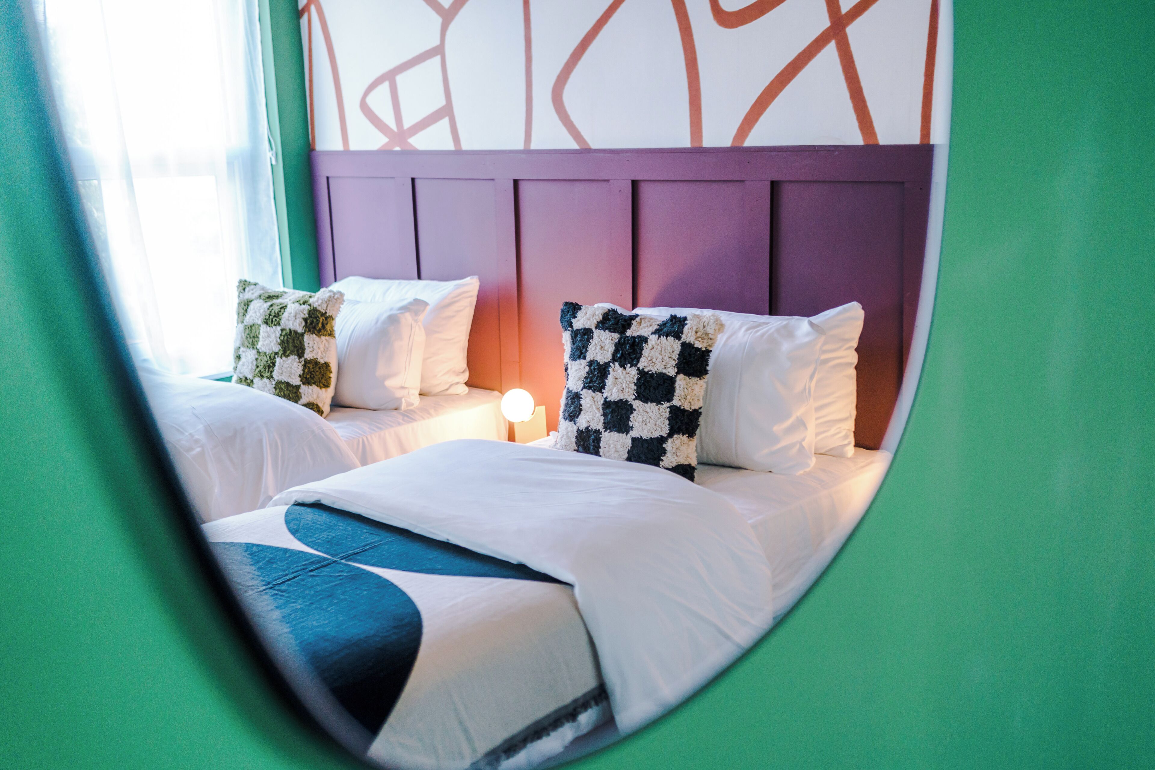 Una habitación con dos camas, cada una con ropa blanca y almohadas a cuadros. Entre las camas, un mesita de noche con una lámpara.