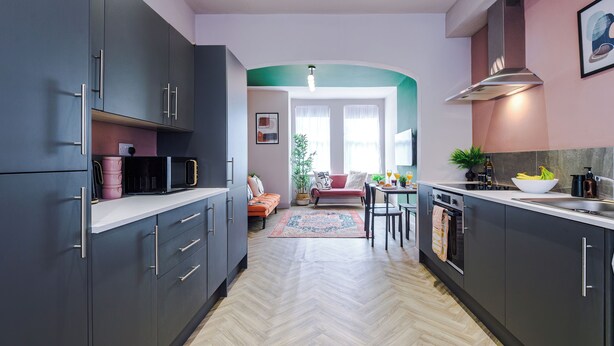 A modern kitchen with dark cabinets, a white countertop, and a herringbone wood floor. There is a dining area with a red sofa and a table with chairs. - Modern 4-Bedroom in Manchester (Salford)