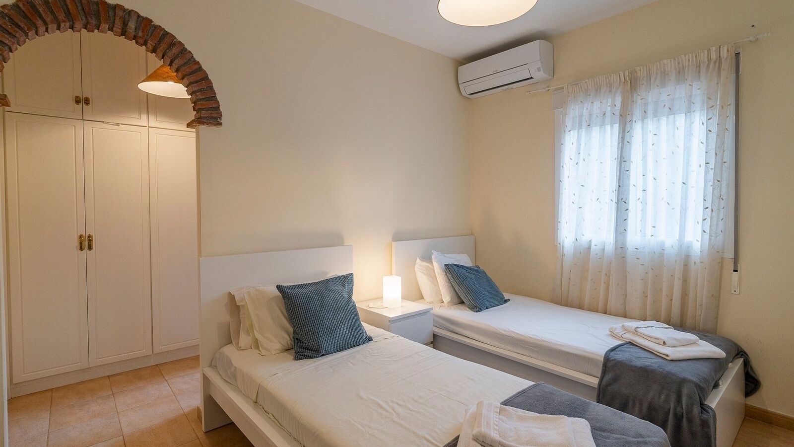 A bedroom with two beds, a wardrobe, an air conditioning unit, and a window with curtains.