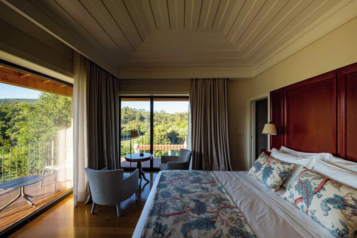 A spacious bedroom with a large bed, a balcony with a table and chairs, and a view of a forested area.