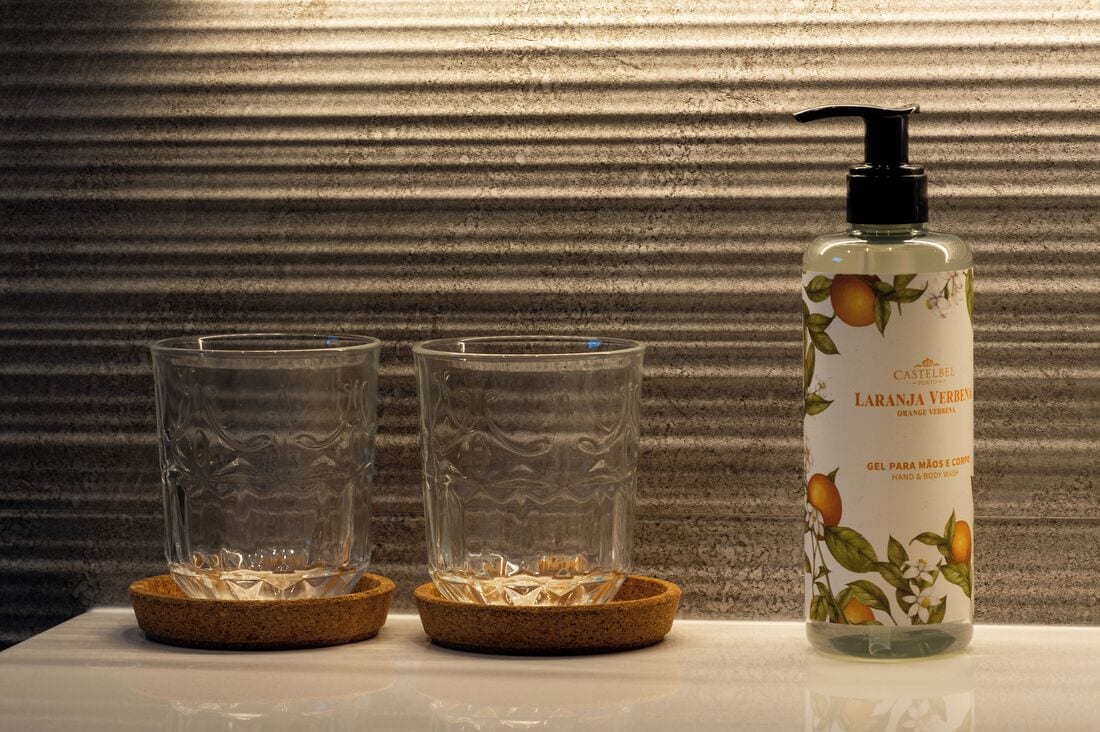 Una botella de jabón de manos y cuerpo Castelbel con dispensador, dos vasos vacíos sobre una mesa con posavasos de corcho.