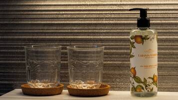 Una botella de jabón de manos y cuerpo Castelbel con dispensador, dos vasos vacíos sobre una mesa con posavasos de corcho.