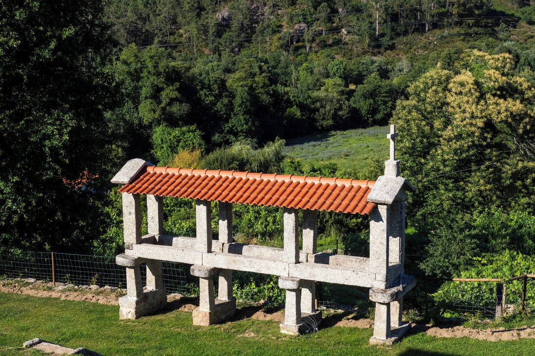 Un banc de pedra amb teulada de rajola en una zona verda, amb arbres al fons.