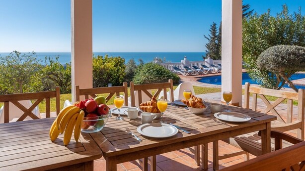 A wooden table set for breakfast with fruits, croissants, and orange juice, overlooking a pool and ocean. - Finca Kriek Rincón de la Victoria by Ruralidays (Málaga)