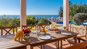 Mesa de madera preparada para el desayuno con frutas, croissants y jugo de naranja, con vista a una piscina y al océano.