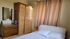 Una cama bien hecha con sábanas blancas y una toalla doblada, un armario de madera con varios compartimentos, y un espejo grande en la pared.