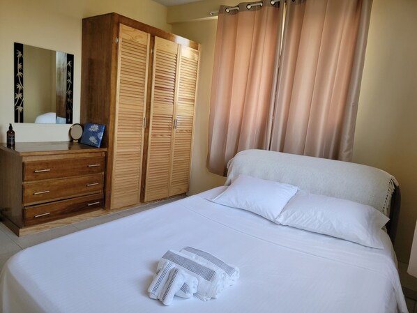 A neatly made bed with white linens and a folded towel, a wooden wardrobe with multiple compartments, and a large mirror on the wall.
