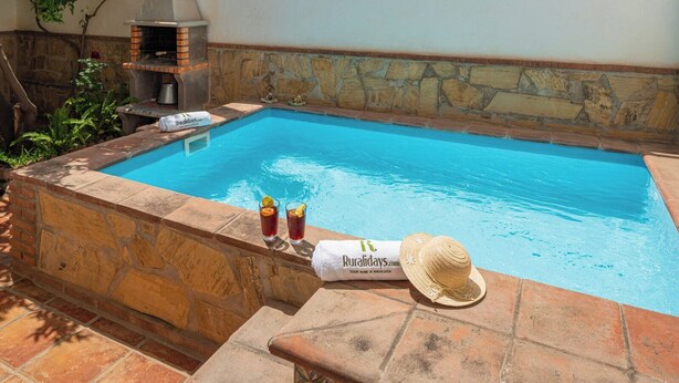 Une piscine bordée d’un mur en pierre, deux boissons et une serviette avec un chapeau.