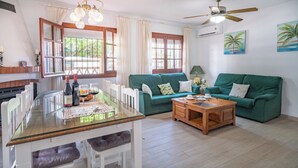 Un salon spacieux avec une table à manger en verre, deux canapés verts, une table basse en bois et un ventilateur de plafond.