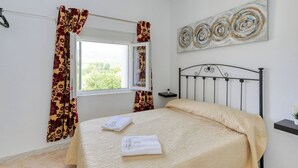 Una cama bien hecha con una colcha color beige y dos toallas dobladas, una ventana con cortinas, una obra de arte colgada en la pared y una mesita de noche con una planta.