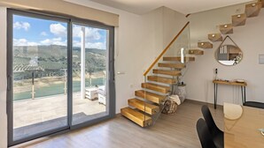 Un interior moderno con una escalera de madera, una zona de comedor y un gran ventanal corredizo que ofrece vistas al paisaje.