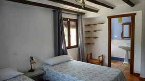 Une chambre avec deux lits, une chaise en bois, un bureau avec une lampe, et une salle de bain visible à travers une porte ouverte.