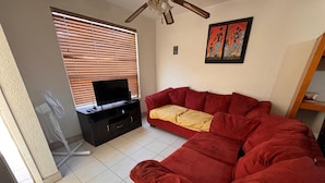 A living room with a red sofa, a television on a black stand, a ceiling fan, and a window with blinds. - Consulado casa de Lujo (Ciudad Juárez)