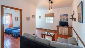 A living room with a sofa, a TV, and a small table with a plant. There are two beds in the adjacent room, a window with blinds, and a painting on the wall. - Zona tranquila cerca de playa Conil de la Frontera by Ruralidays (Cádiz)