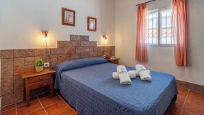 A hotel room with a stone headboard, a blue quilted bedspread, two rolled towels with a logo, a nightstand with a plant, and a window with striped curtains. - Zona tranquila cerca de playa Conil de la Frontera by Ruralidays (Cádiz)