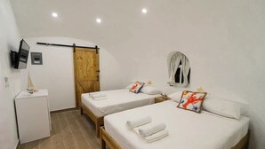 Una habitación pequeña y limpia con dos camas individuales, una puerta de madera, un escritorio pequeño y un televisor montado en la pared.