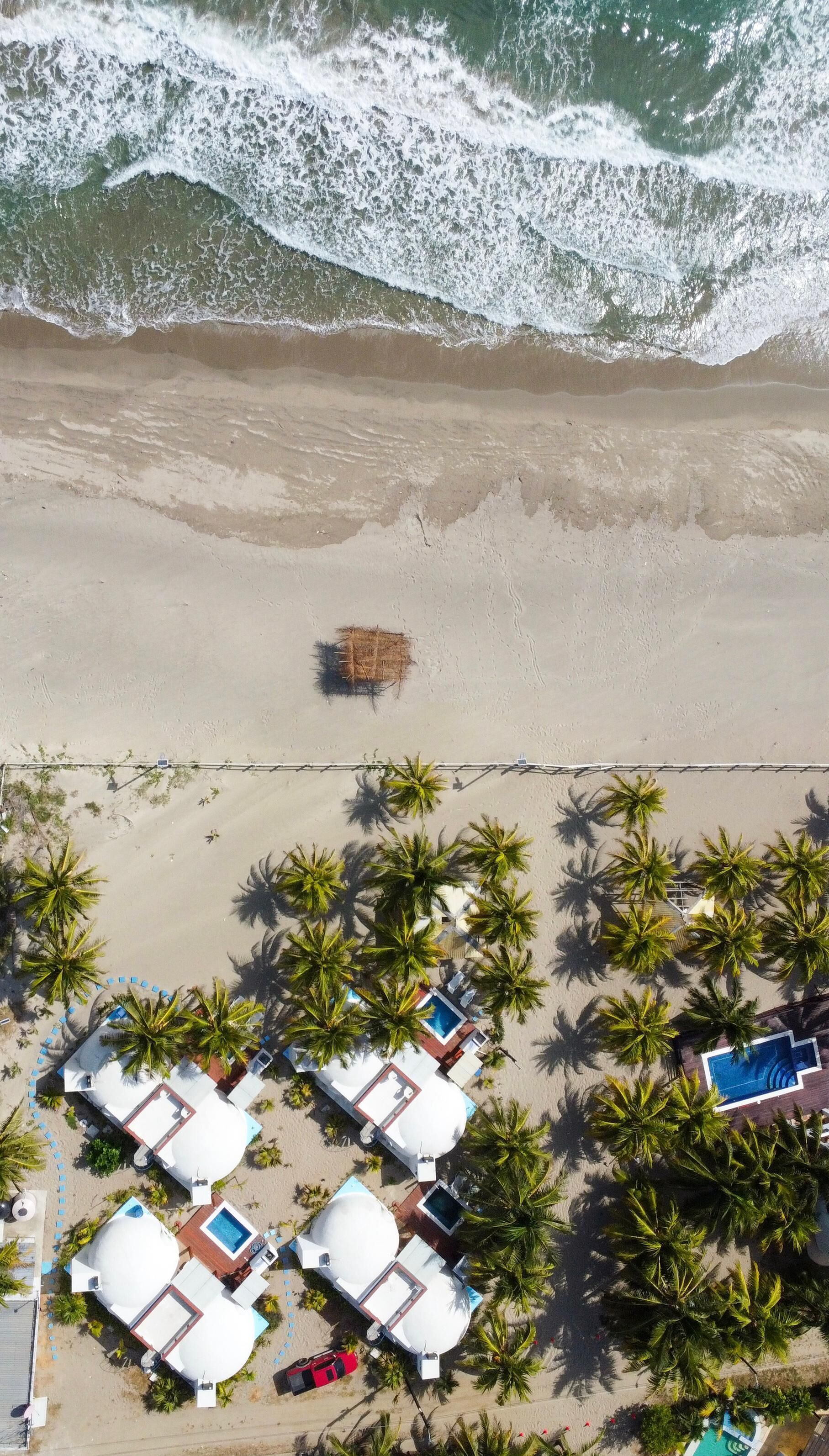Vista aérea de un complejo turístico en la playa con varias cabañas, una piscina y una zona de playa con palmeras.