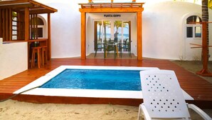 Un área de piscina con terraza de madera, una silla de descanso blanca y una pérgola de madera con una mesa y sillas.