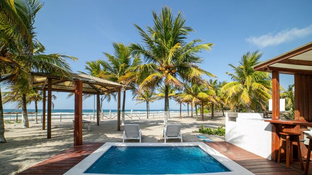 A pool area with a wooden deck, two white lounge chairs, and a view of the beach and ocean.