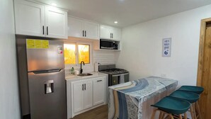 Cocina compacta con gabinetes blancos, refrigerador de acero inoxidable, microondas y estufa. Incluye dos taburetes de bar y una pequeña isla con mantel estampado.