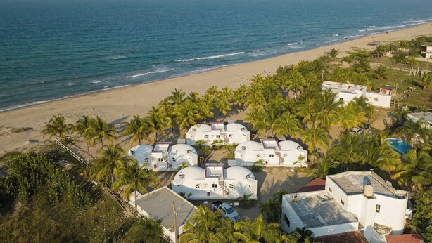 Una zona costera con edificios de forma abovedada blanca rodeada de palmeras, con vistas a la playa y el océano.