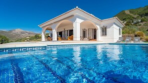 Una villa blanca con piscina, que cuenta con un porche cubierto con sillas y vistas a las montañas.