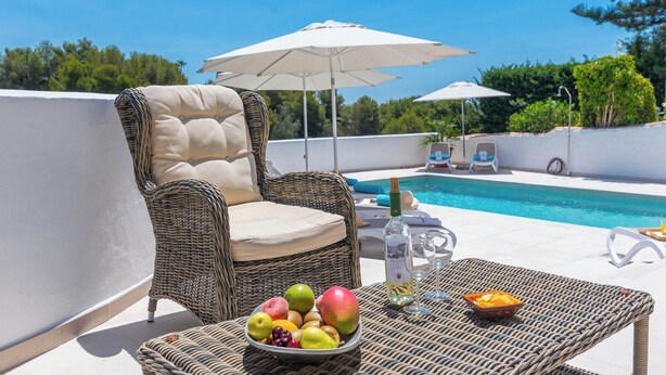 A wicker lounge chair with a cushion, a table with a fruit bowl and a bottle, and a pool in the background. - Villa Higuera Nerja by Ruralidays (Málaga)