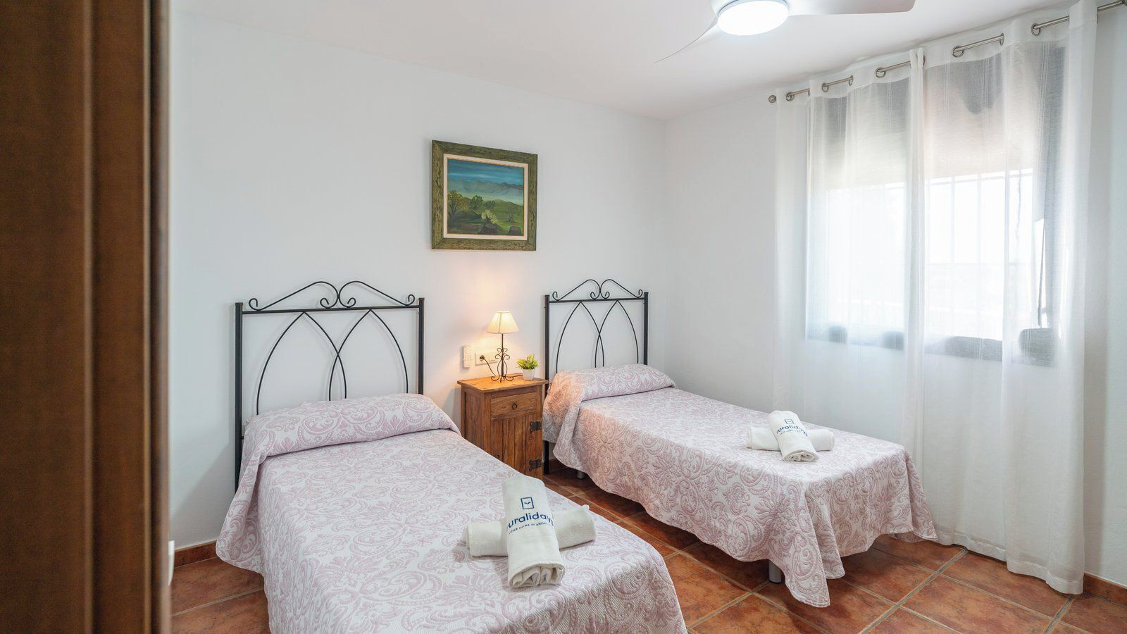 A room with two beds, each with a patterned bedspread and pillows, a nightstand with a lamp, and a painting on the wall.