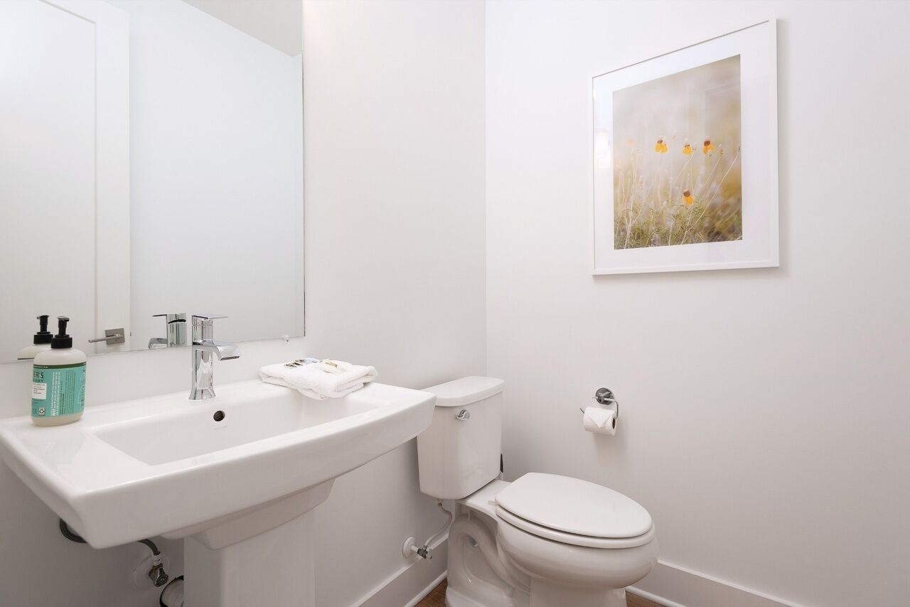 A modern bathroom with a white sink, mirror, and toilet. There is a framed picture on the wall and a bottle of hand soap on the sink.