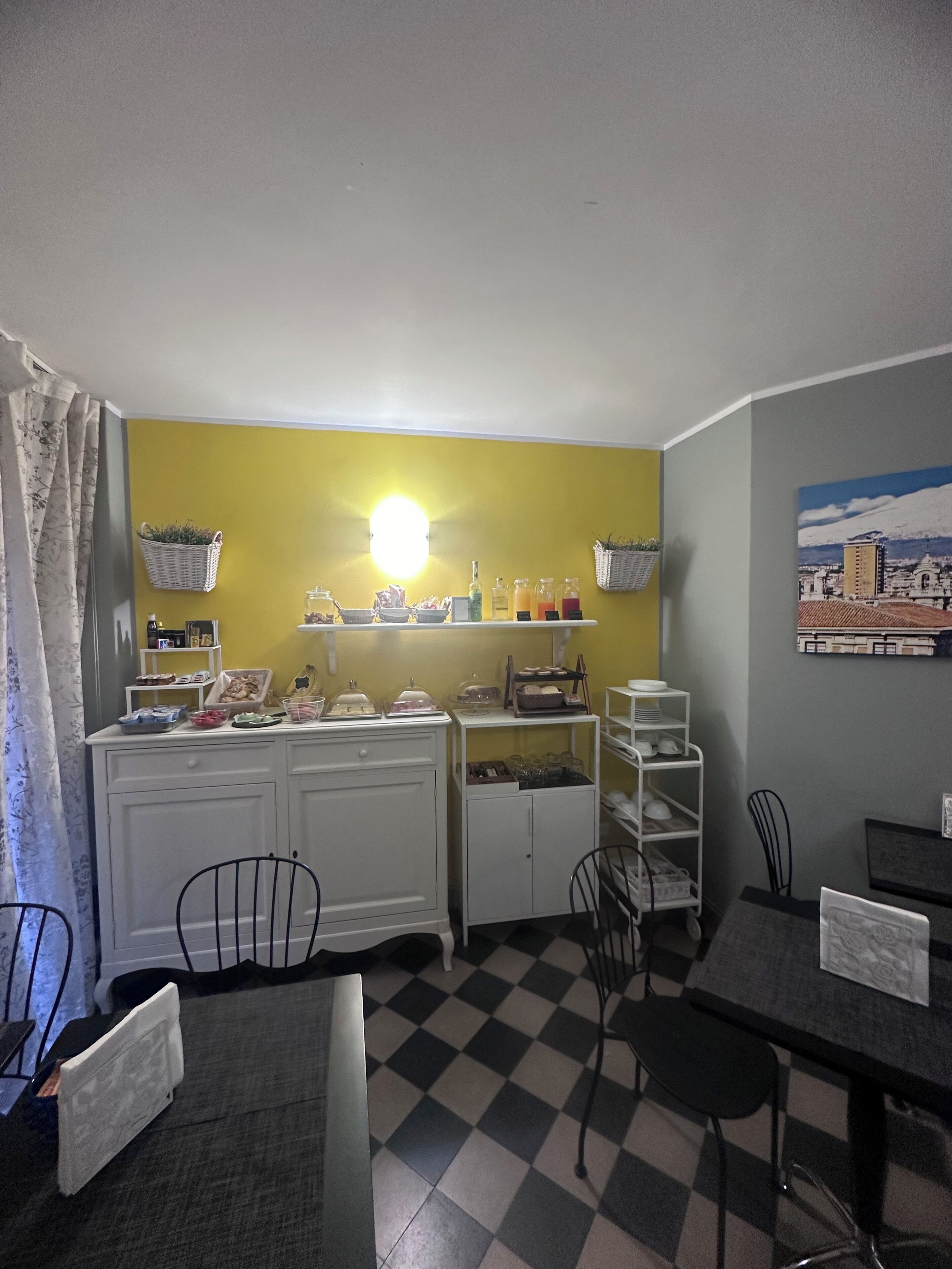 Una camera d'albergo compatta con pavimento a scacchi, un'area pranzo con tavolo e sedie, un angolo cottura con mobiletti e scaffali bianchi e un quadro raffigurante un panorama urbano.