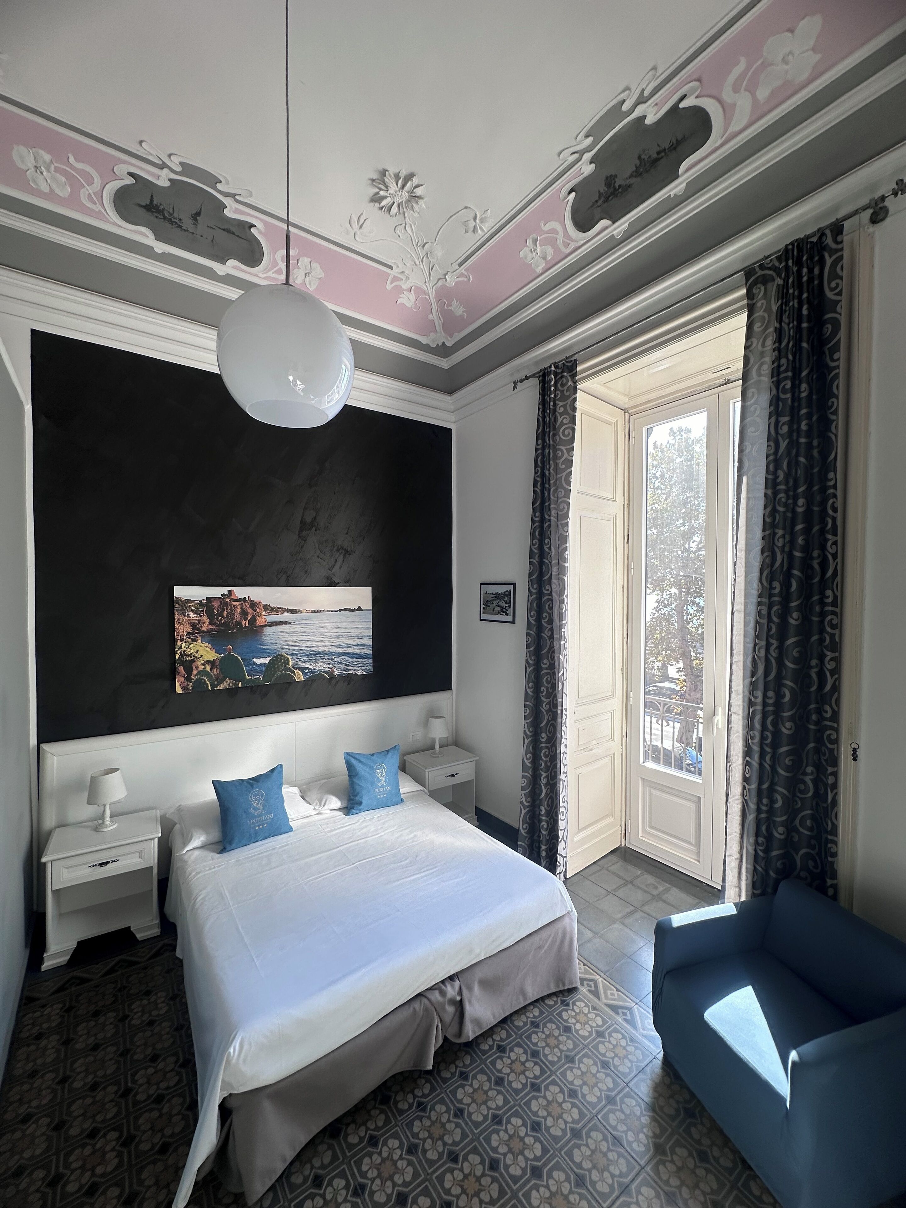 Une chambre à coucher avec un grand lit, une table de chevet, une chaise bleue, un luminaire au plafond, une fenêtre avec des rideaux et un tableau accroché au mur.