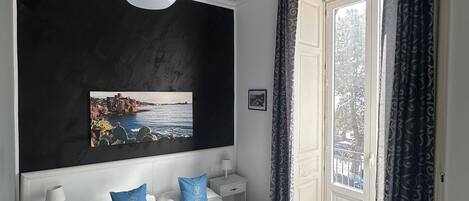 Une chambre à coucher avec un grand lit, une table de chevet, une chaise bleue, un luminaire au plafond, une fenêtre avec des rideaux et un tableau accroché au mur.
