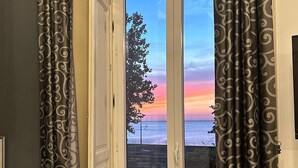 A room with a large window featuring a view of a sunset over the ocean, a white bathtub, and a wooden bench with a basket. - B&B I Puritani (Catania)