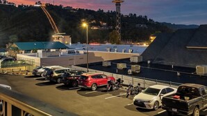 A parking lot with several cars and motorcycles, a building with a flat roof, and a tower in the background.