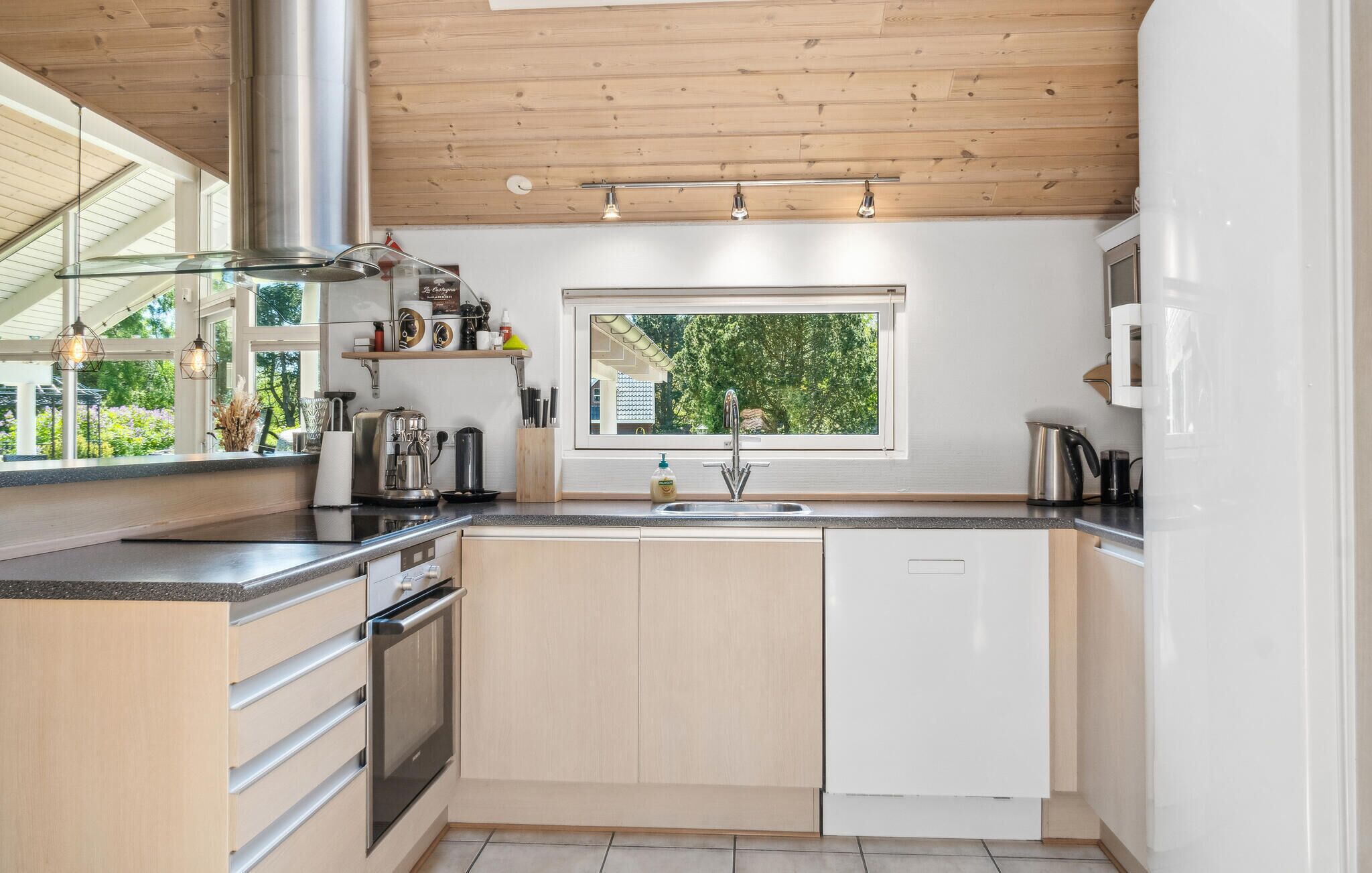 A modern kitchen with a wooden ceiling, a large window, and a view of greenery outside.