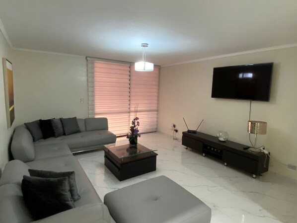 A modern living room with a sectional sofa, a glass-top coffee table, a flat-screen TV mounted on the wall, and a floor lamp.