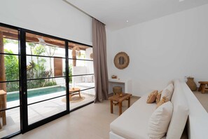 A modern living room with a large sliding glass door leading to a pool area, a white sofa, and a small wooden table. - Villa Camille (Tabanan)