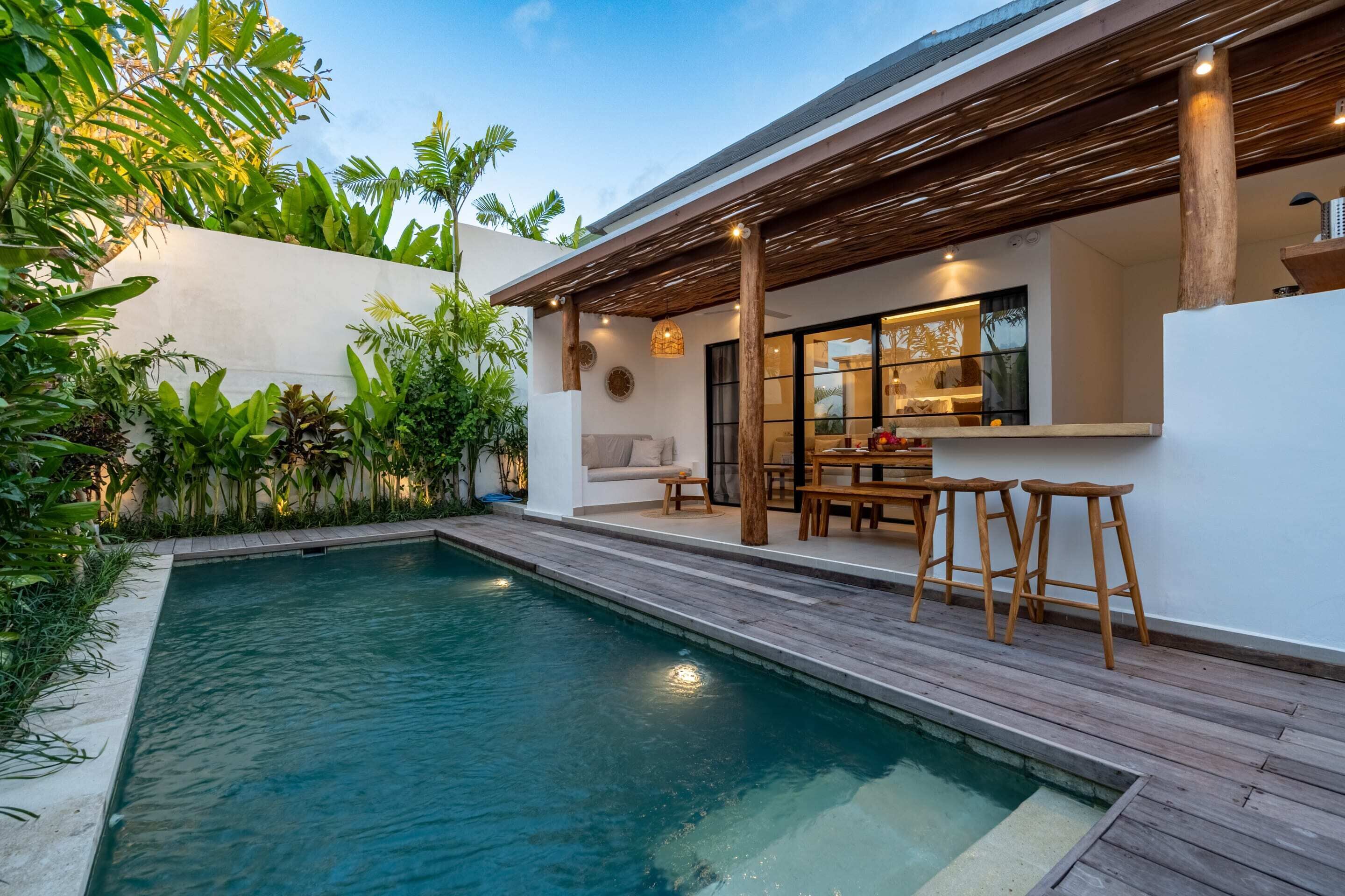 A modern outdoor pool area with a wooden deck, a seating area with a table and chairs, and a bar with stools.