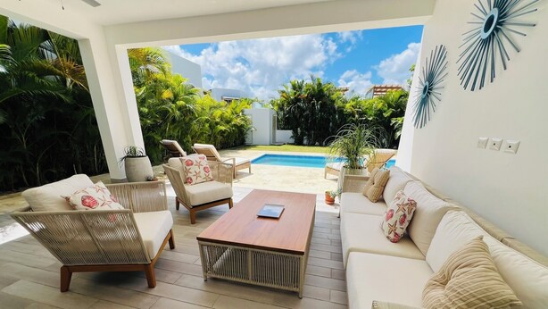 A modern outdoor living area with wicker furniture, a wooden coffee table, and a view of a swimming pool and greenery.