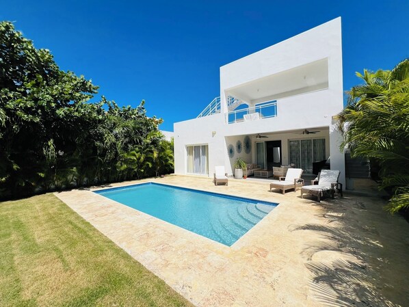 Una casa moderna de dos pisos con piscina, mobiliario exterior y abundante vegetación.