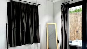A bedroom with a bed, a hanging black curtain, a wooden-framed mirror, and a view of a bamboo fence.
