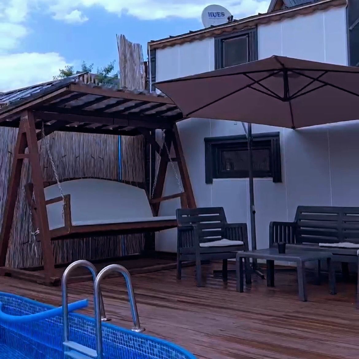 Zona de piscina con una silla colgante de madera, una mesa con sillas y una sombrilla.