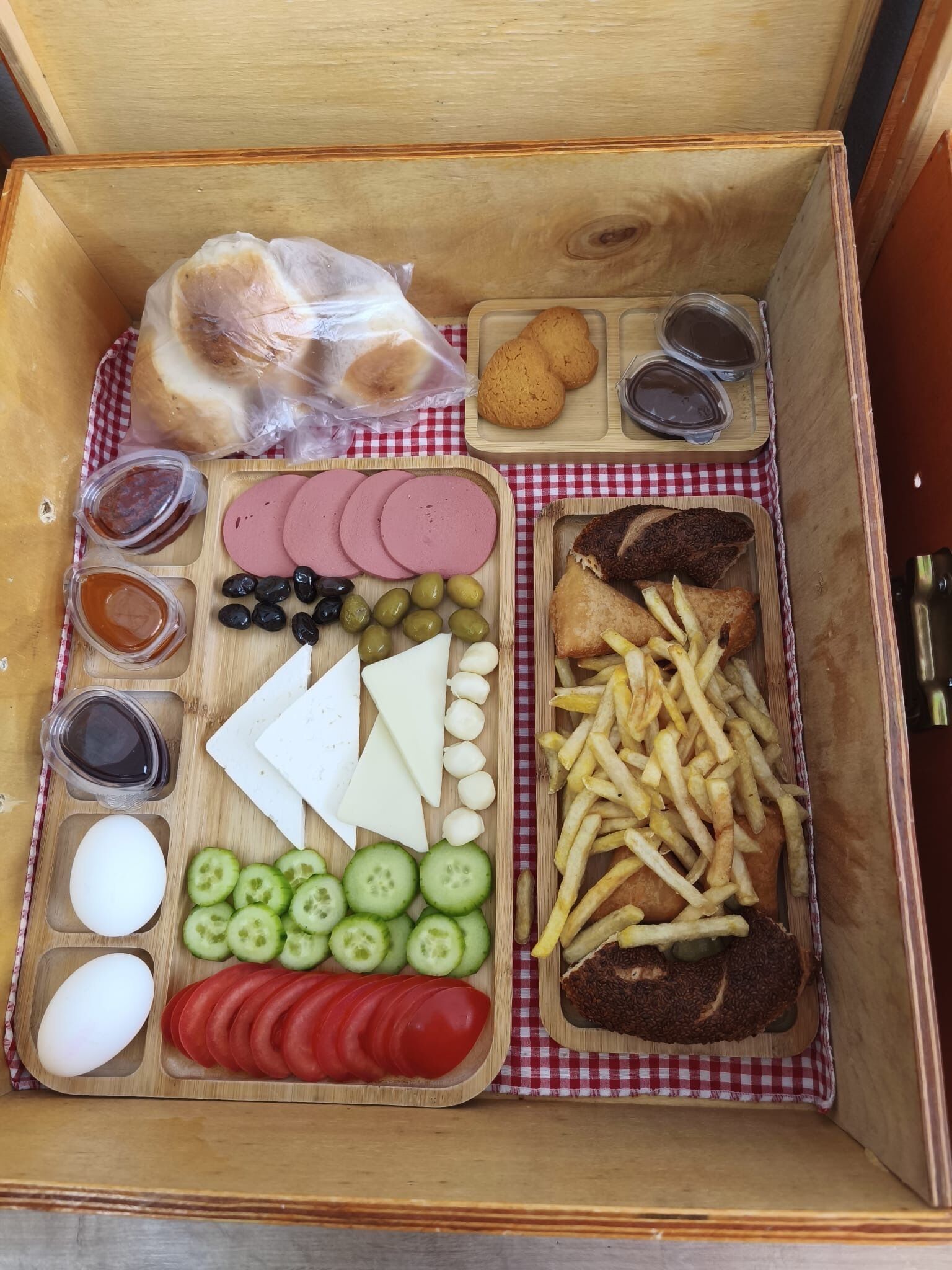 A wooden box containing various food items like sliced meats, cheeses, olives, cucumbers, tomatoes, eggs, bread, and French fries.