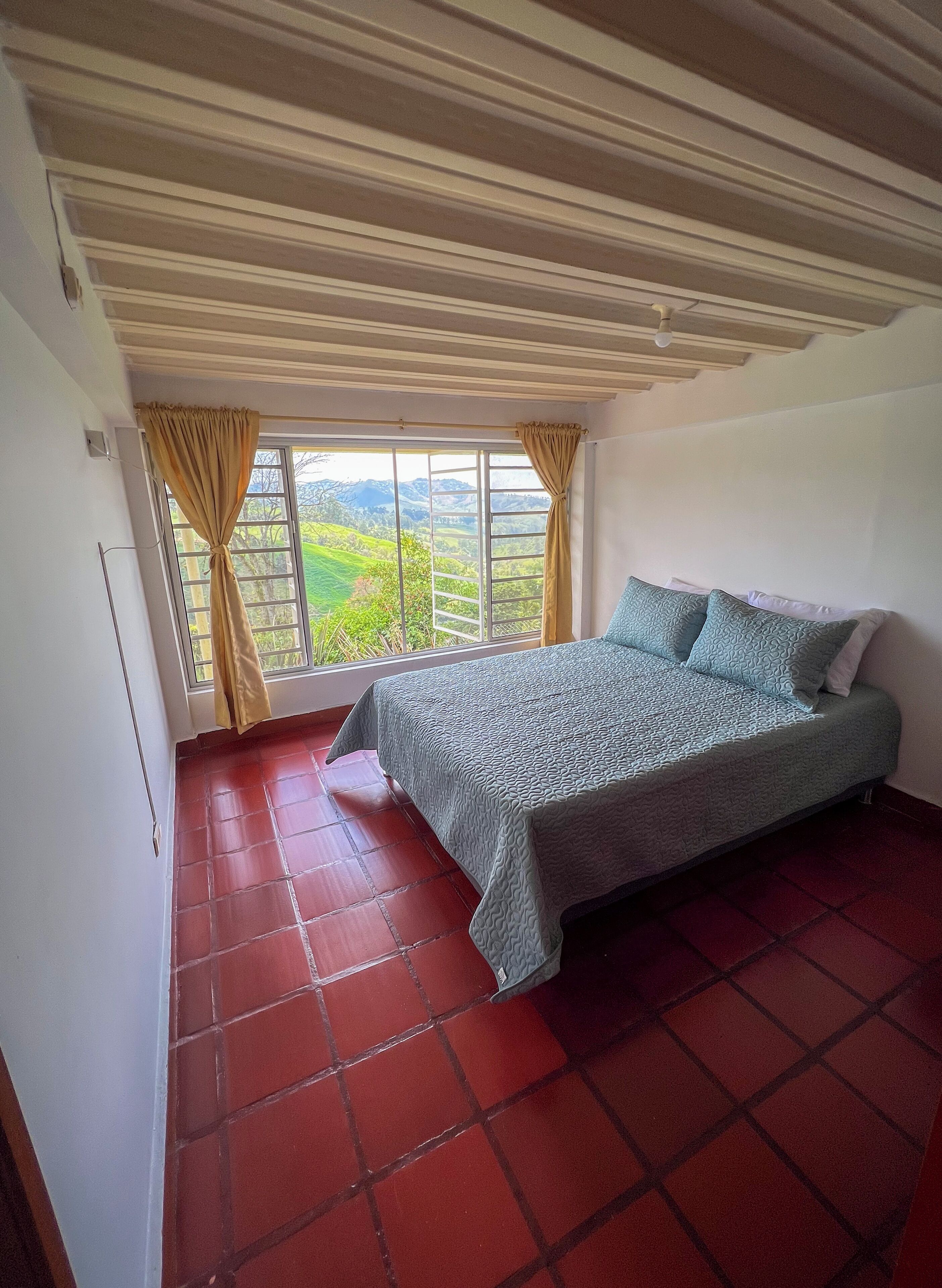 Una habitación con cama, ventana con cortinas y vistas a colinas verdes.