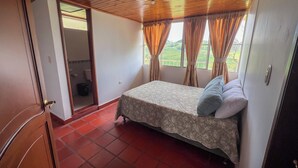 Un dormitori amb un llit, una finestra amb cortines, un sostre de fusta i un paviment de rajoles vermelles.