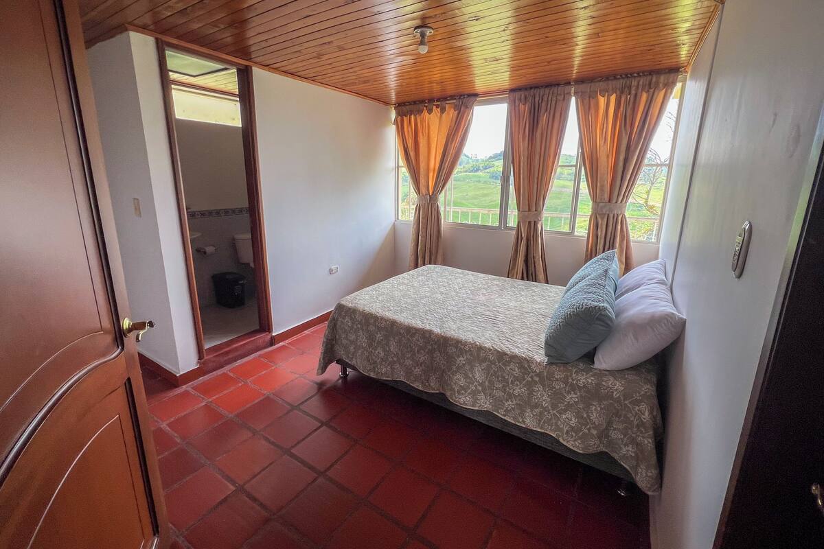 Une chambre à coucher avec un lit, une fenêtre avec des rideaux, un plafond en bois et un sol recouvert de carreaux rouges.