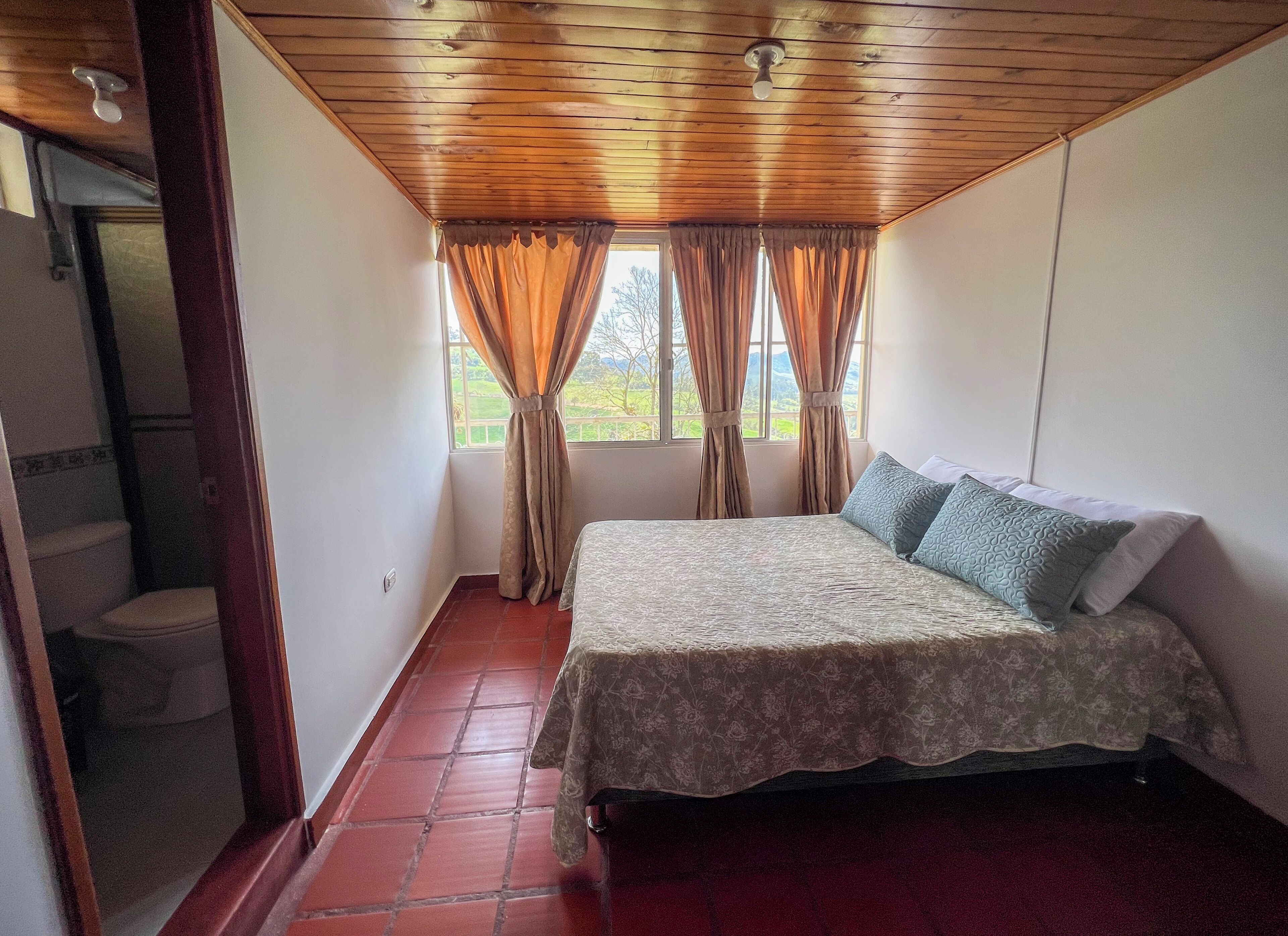 Une chambre à coucher avec un lit, une fenêtre avec des rideaux, un plafond en bois et un sol recouvert de carreaux rouges.